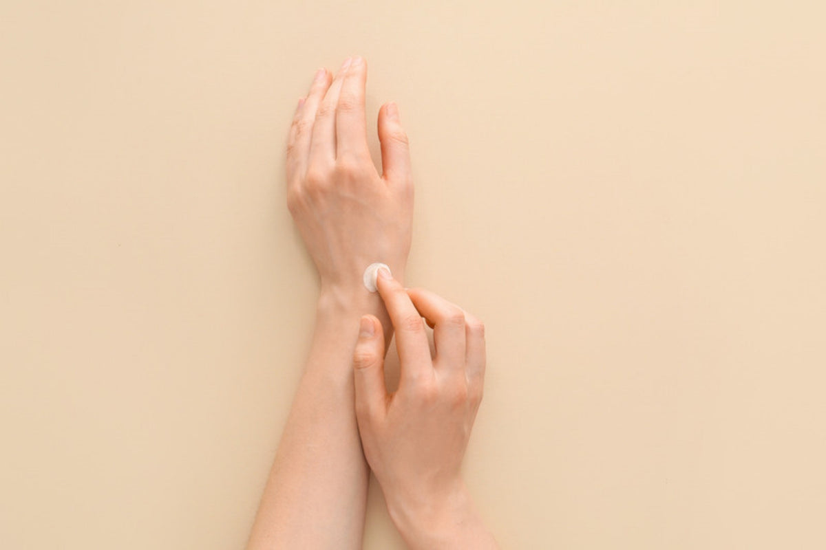 Hands pressing cream into the back of the hand on a warm beige ground in soft light