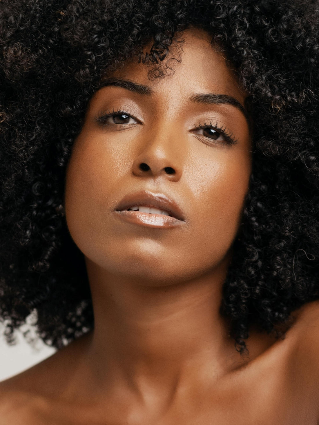 Woman with natural afro hair, quiet direct gaze, warm grey backdrop