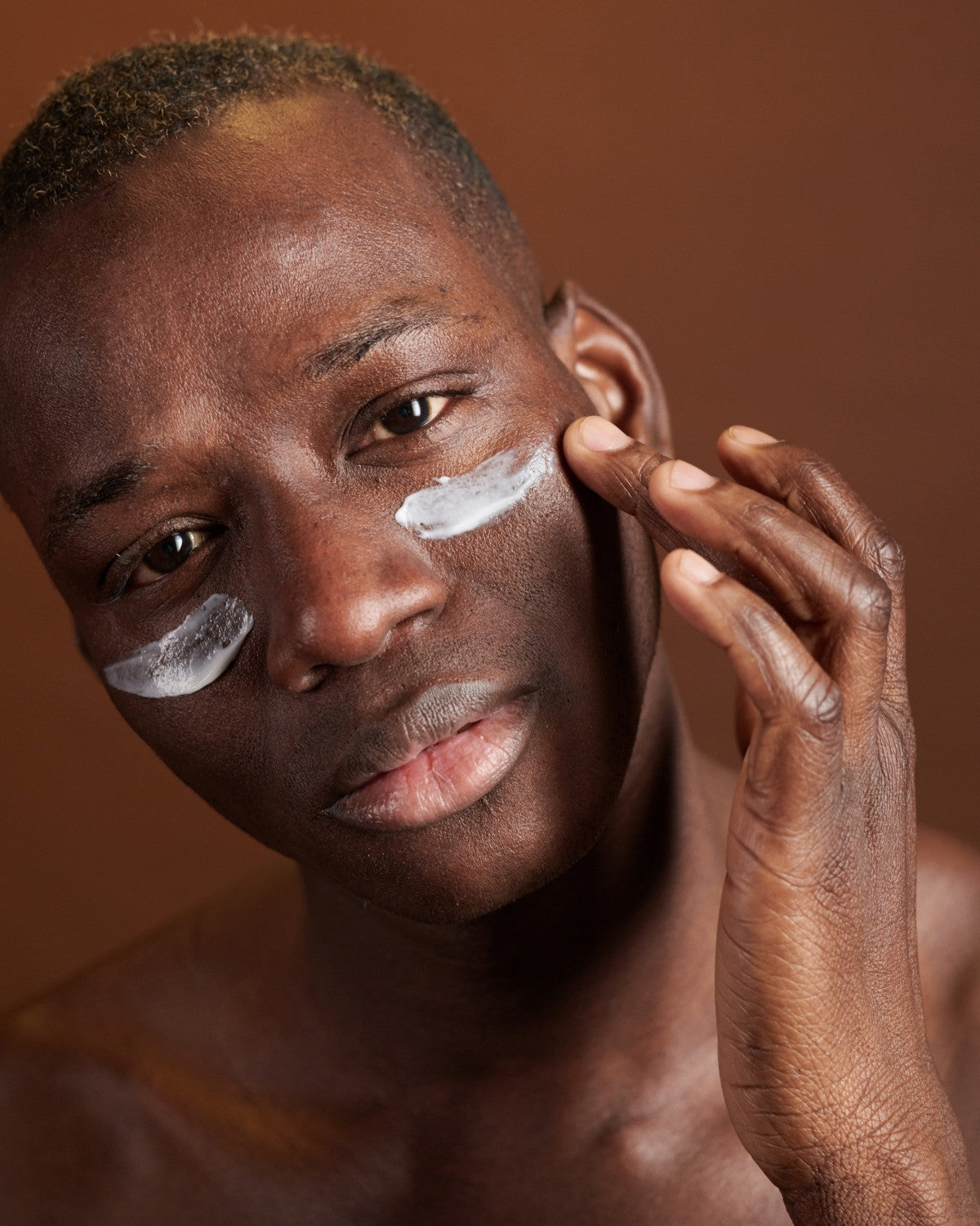 Man pressing cream into his cheek, direct gaze, warm brown studio backdrop